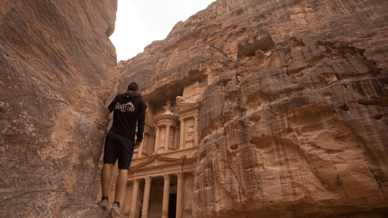 A person in a black jacket stands on a rock ledge, marveling at the ancient stone facade of Al-Khazneh (The Treasury) in Petra, Jordan. With its carvings etched into towering cliffs amidst rugged, reddish-brown terrain, one might wonder: Do I need a visa for Jordan to explore such wonders?.
