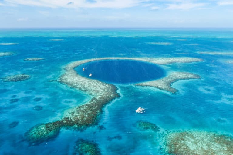 Aerial view of the Great Blue Hole in Belize showing a large dark blue circular sinkhole surrounded by lighter turquoise coral reefs and several small boats nearby on the water