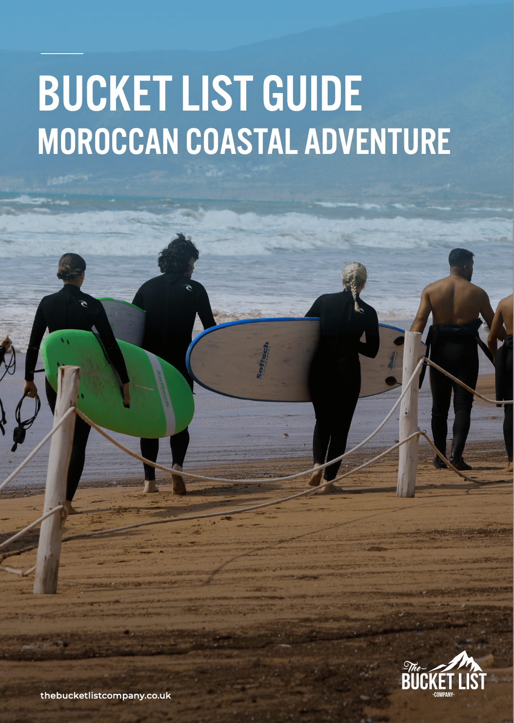 Five surfers in wetsuits stride across a sandy beach towards the ocean carrying surfboards of various colors The sky is partly cloudy and waves crash in the distance A sign reads "Bucket List Guide: Morocco Adventure Ride the Coastal Waves