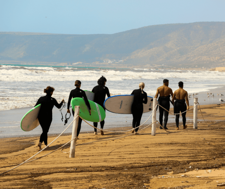 A group of six wetsuit clad surfers carries surfboards along a sandy beach at a surf yoga retreat in Morocco Ocean waves crash nearby and posts with ropes separate the beach from the dunes Hills rise in the background under a clear sky