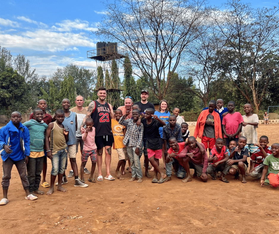 A group of children and adults pose together outdoors on the dirt ground near the Kilimanjaro Marangu Route with trees a water tower and a blue sky in the background Everyone appears happy and cheerful